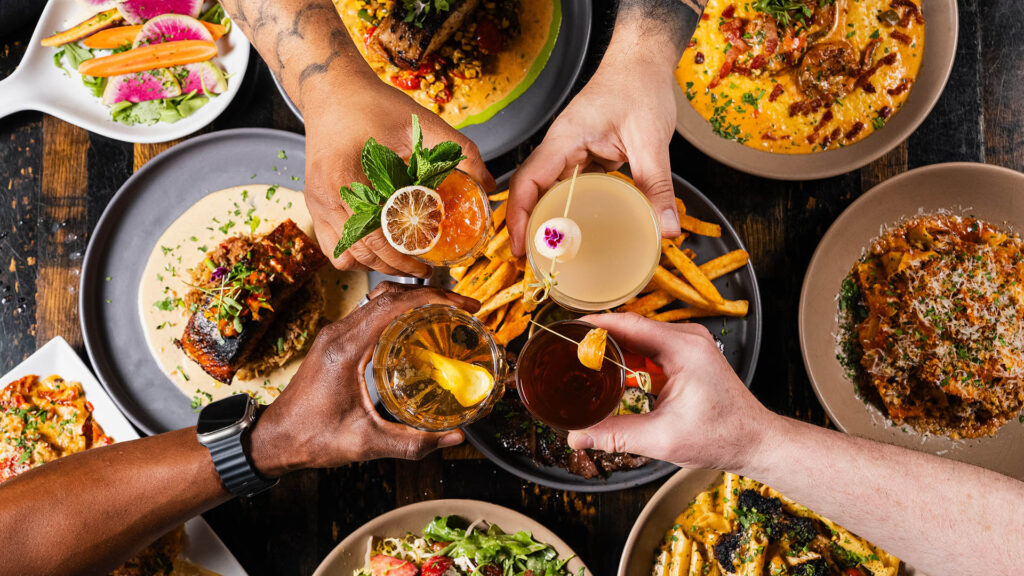 People holding cocktails over several different entrees and appetizers at Bottled in Bond Cocktail Parlour and Kitchen